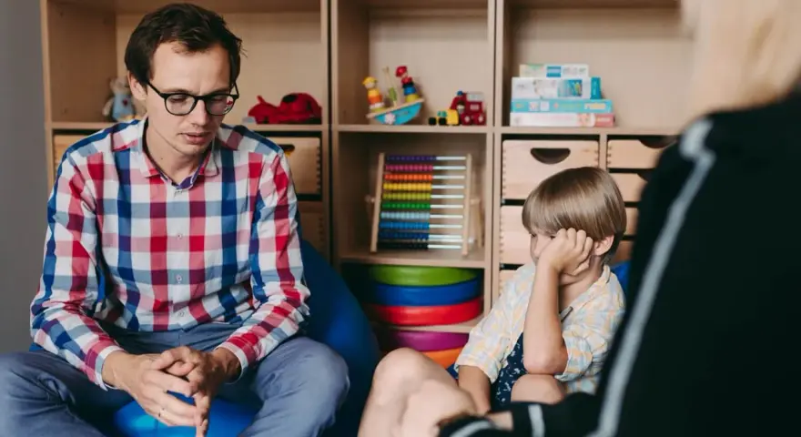 Terapeuta rozmawia z dzieckiem, ćwicząc teorię umysłu. W tle zabawki i pomoce edukacyjne.