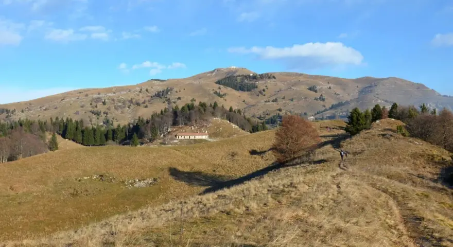 Sentieri facili sul Monte Grappa, con un escursionista che percorre un crinale erboso verso una casa isolata e la cima della montagna in lontananza.