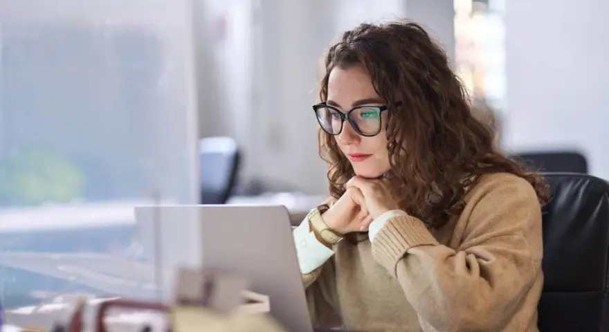 Donna con occhiali lavora al laptop, pensierosa. Potrebbe essere una dei nomadi digitali chi sono, che lavora da remoto.