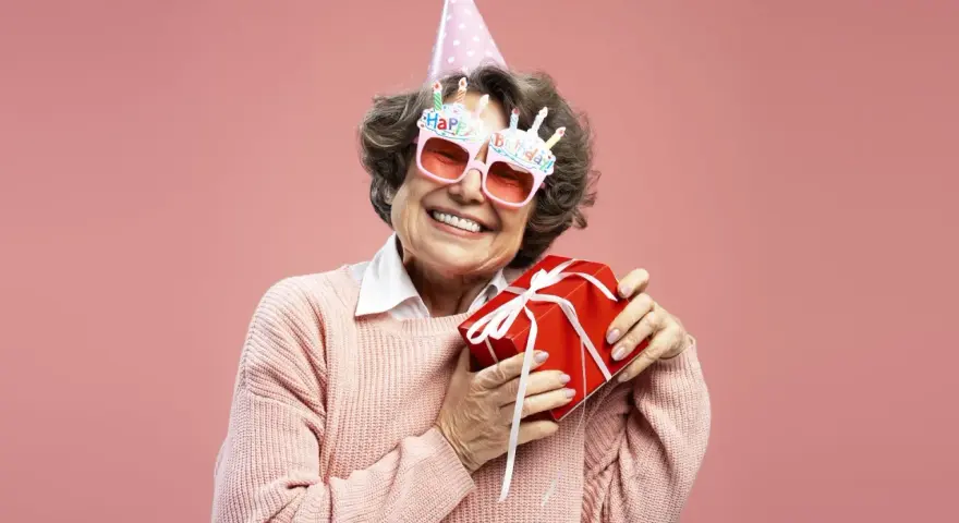 Uśmiechnięta starsza pani w okularach z napisem "Happy Birthday" i w czapce urodzinowej trzyma prezent. To dowcipne podziękowania za życzenia!