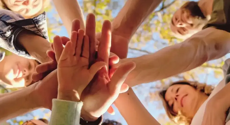Grupa ludzi złącza dłonie w geście jedności i altruizmu. Uśmiechnięte twarze patrzą w górę, symbolizując wspólny cel i wsparcie.