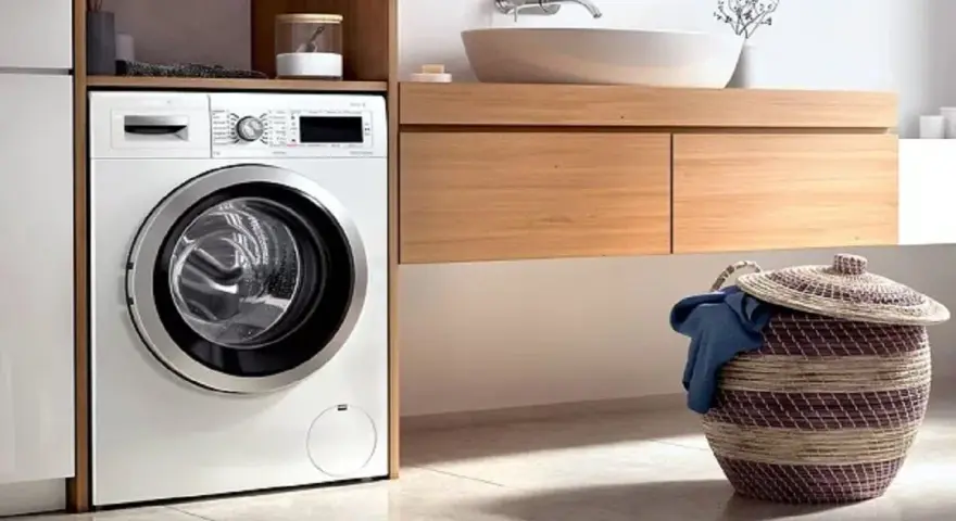 Lavadora blanca moderna lista para instalar en un baño con cesta de ropa sucia.