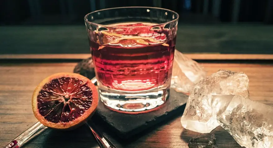 Ein Glas mit einem roten Drink, garniert mit Orangenschale, neben einer Blutorange und Eiswürfeln. Das ist Campari, ein italienischer Aperitif.