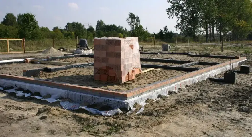 Stan zero budynku: fundamenty z cegły i betonu, piasek, folia izolacyjna, stos cegieł pod przykryciem.