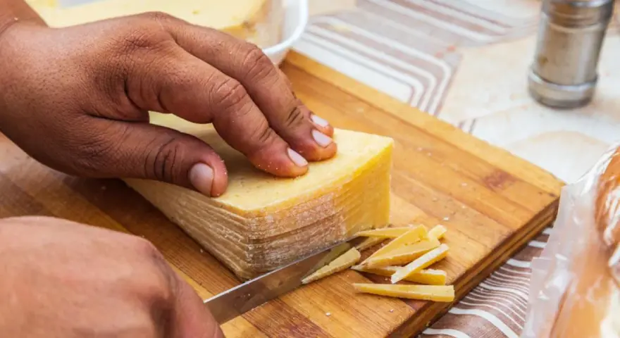 Mano cortando un bloque de queso amarillo en finas láminas. La corteza del queso se ve firme, indicando que se puede comer.