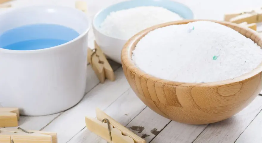 Bol en bois rempli de lessive en poudre maison, à côté d'une tasse de liquide bleu et de pinces à linge en bois.