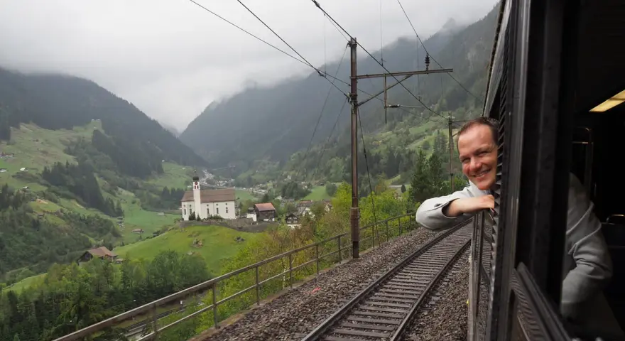 A smiling man leans out the window of the Gotthard Panorama Express, enjoying the stunning Swiss Alps scenery with a church nestled in the valley below.