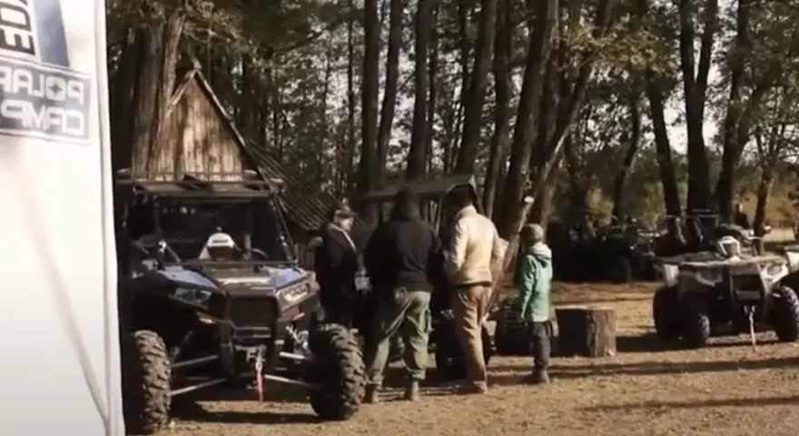 Grupa ludzi przy pojazdach terenowych podczas Polaris camping. W tle las i drewniana chata.