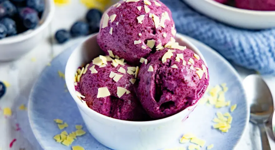 Drei Kugeln Blaubeer Eis, garniert mit weißen Schokostückchen, in einer weißen Schale auf einem blauen Teller.