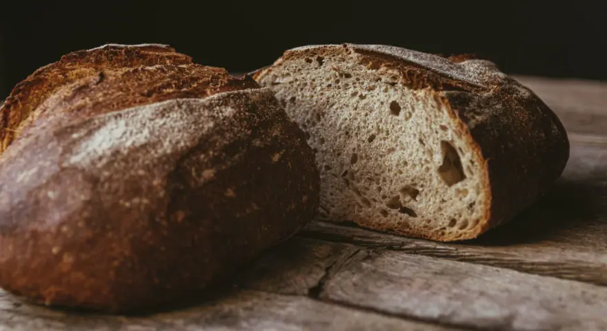 Ein rustikales, aufgeschnittenes sauerteigbrot ohne kneten liegt auf einer Holzoberfläche. Die Kruste ist dunkelbraun und knusprig, das Innere luftig und porös.