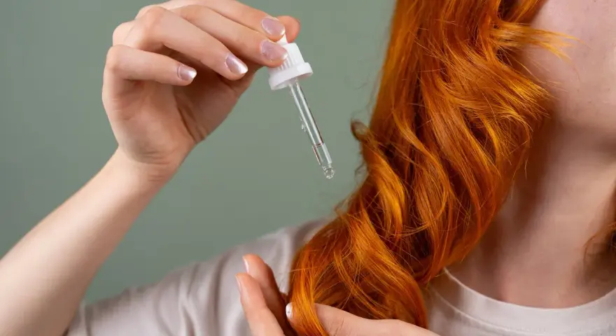 Dłoń z pipetką z olejkiem aplikuje pielęgnację na rude, falowane włosy. Dowiedz się, jak często olejować włosy dla najlepszych efektów.