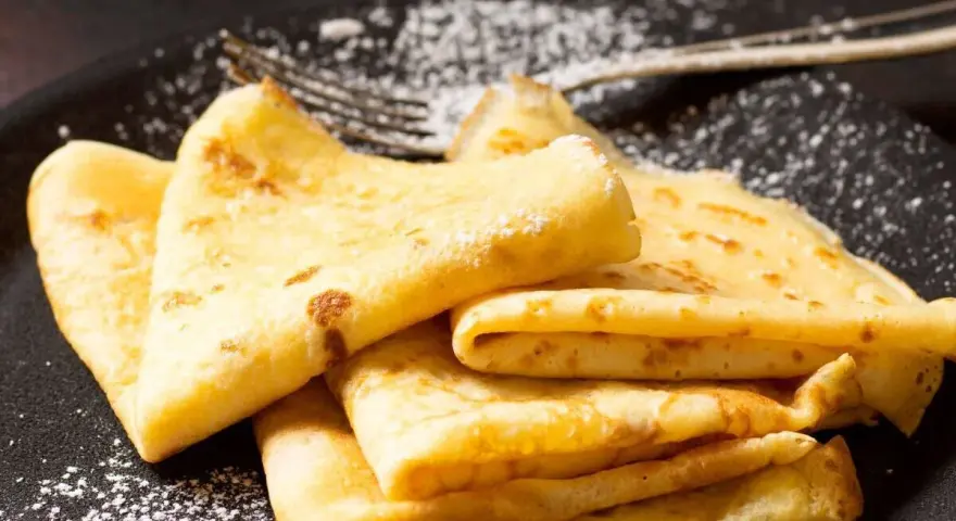 Deliciosos crepe en francés, doblados y espolvoreados con azúcar glas sobre un plato negro.