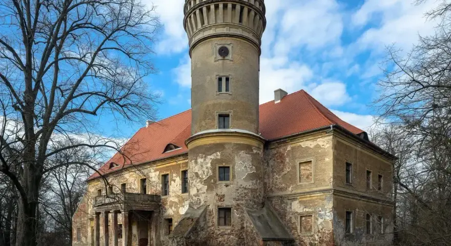 Zniszczony pałac w Osetnie z charakterystyczną wieżą i czerwoną dachówką, otoczony drzewami.
