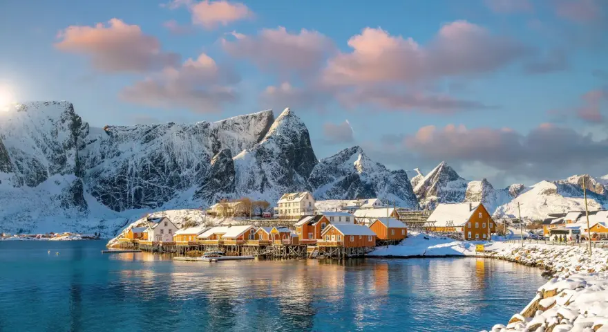 Snow-covered mountains frame a picturesque village with orange and white houses on stilts over the water, a scene from one of Europe's best winter destinations.