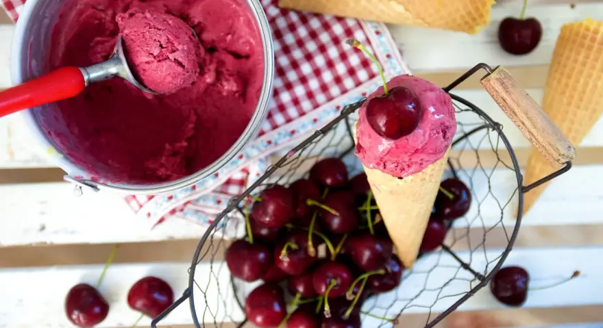 Ein Eisbecher mit Kirscheis, eine Kugel in einer Waffel, frische Kirschen und leere Waffeln.