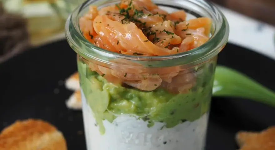 Leckerer Lachs Avocado Vorspeise im Glas, geschichtet mit cremiger Sauce und garniert mit Dill. Dazu geröstete Brotscheiben.