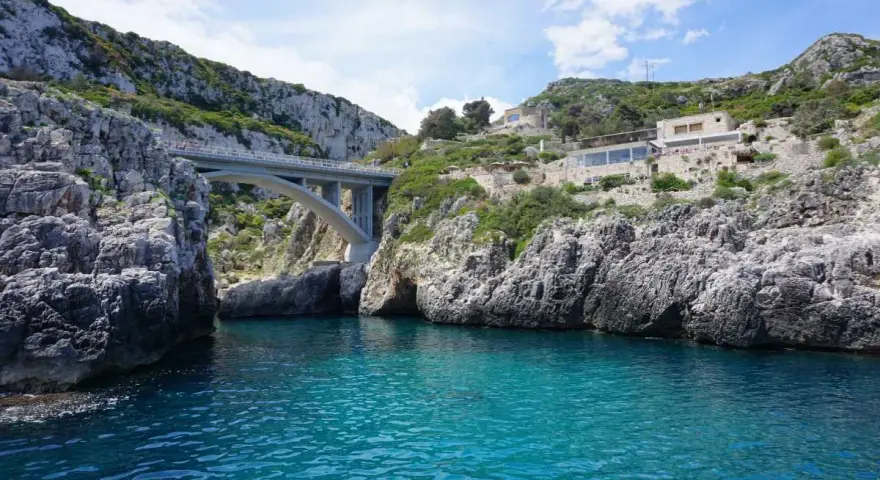 Ponte moderno su un mare cristallino, incastonato tra scogliere rocciose. Un luogo magico dove si incontrano i due mari in Puglia, con un ristorante panoramico sulla collina.