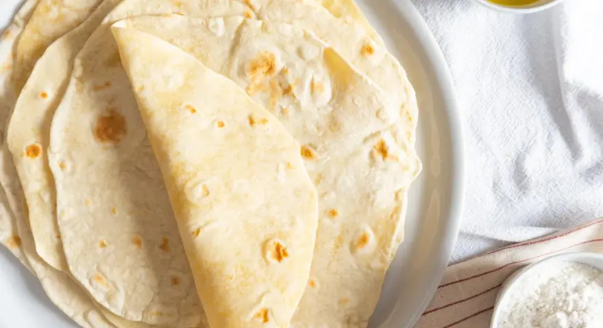 Piatto di piadine senza strutto, morbide e dorate, pronte per essere farcite. Accanto, una ciotolina d'olio e una di farina.