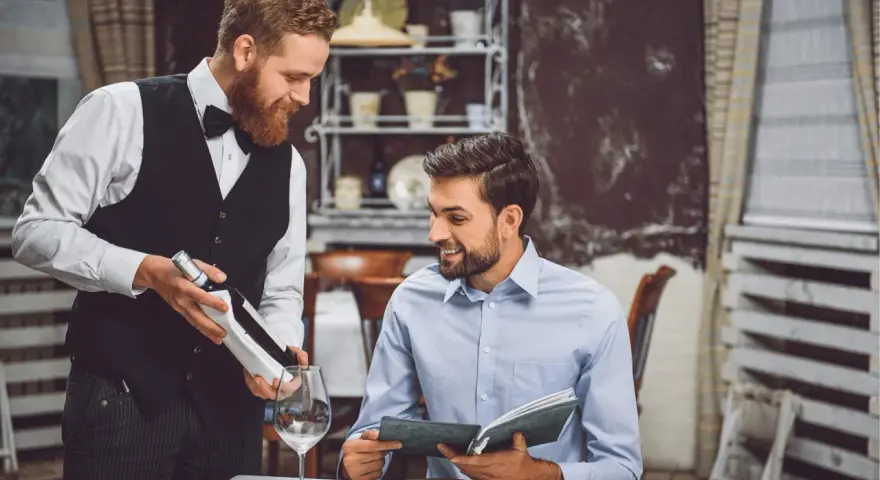 Camarero atento presenta una botella de vino a un cliente que revisa el menú. Demuestra las cualidades de un camarero: servicio, amabilidad y conocimiento.