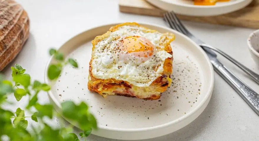 Pyszny croque madame z jajkiem sadzonym na wierzchu, podany na talerzu z pieprzem i ziołami.