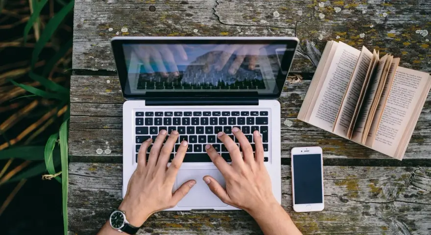Ręce piszą CV funkcjonalne na laptopie, obok leży otwarta książka i telefon.