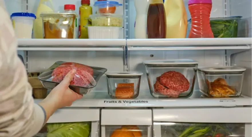 Mano colocando carne picada envasada en un refrigerador lleno de alimentos.