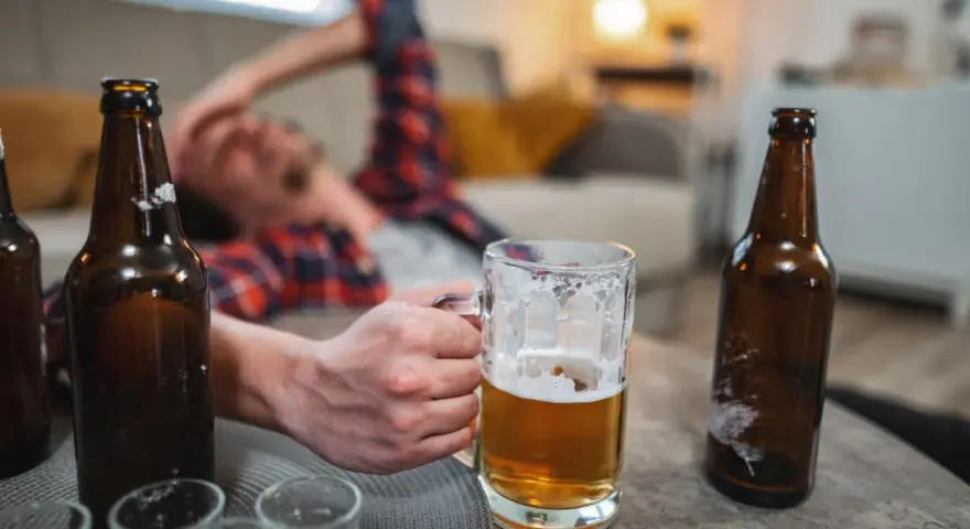 Mężczyzna leży na kanapie, trzymając kufel piwa. Wokół puste butelki, sugerujące nagły spadek tolerancji na alkohol.