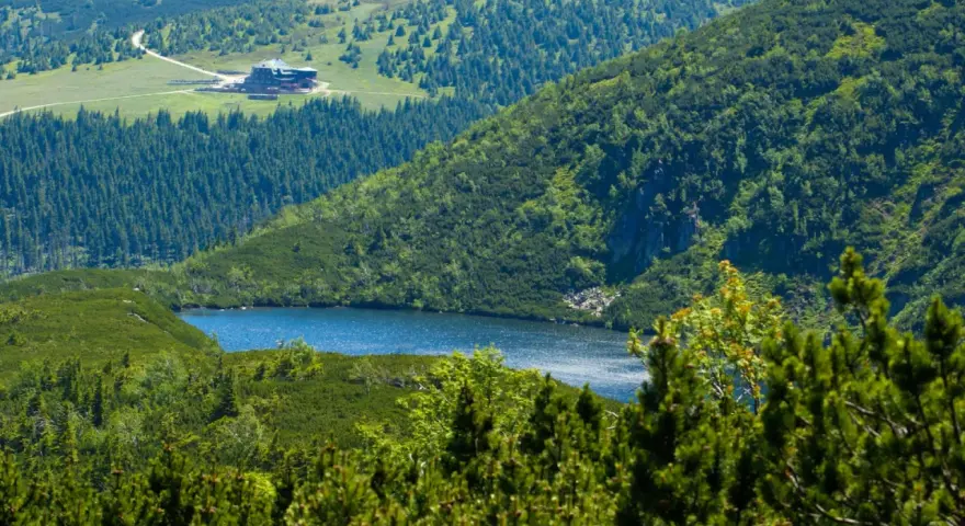 Widok na górskie jezioro i schronisko Samotnia w Karkonoszach. Otoczone zielenią, lasem i skalistymi zboczami.