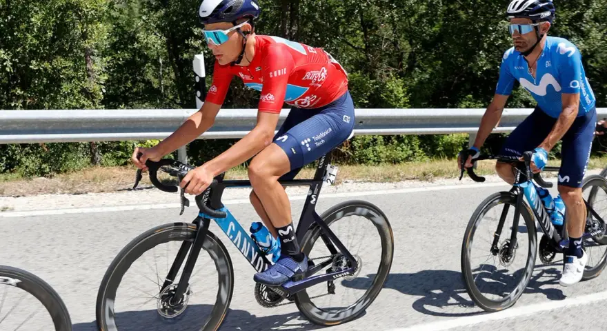 Cyclists in red and blue vuelta jerseys race on a sunny road, with lush green trees lining the background.