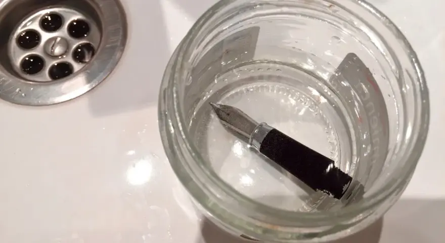 Pluma estilográfica sumergida en un frasco de vidrio con agua, junto a un desagüe. Un paso para aprender como limpiar una pluma.