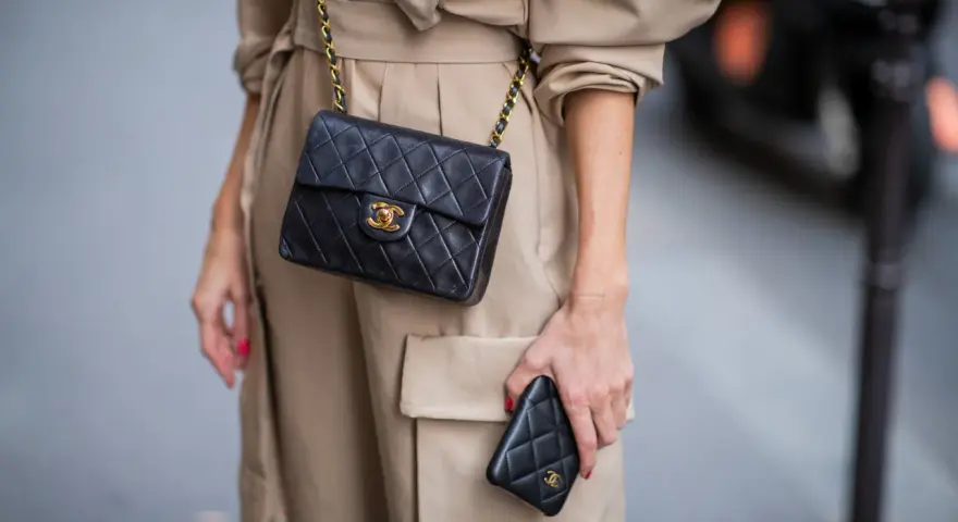 Elegancka kobieta w beżowym płaszczu prezentuje klasyczną, pikowaną torebkę Chanel vintage z złotym łańcuszkiem i logo.