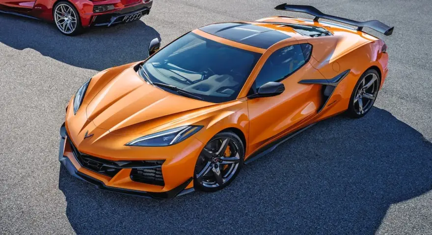 Pomarańczowy Chevrolet Corvette C8 z czarnym dachem i spojlerem na torze wyścigowym. Obok widać fragment czerwonego samochodu.