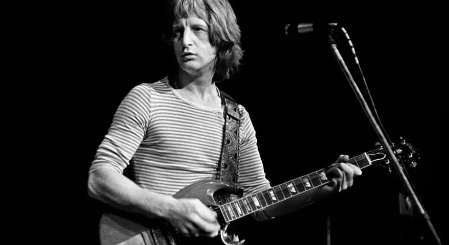 Pete Ham, mit gestreiftem Shirt und Gitarre, blickt konzentriert auf die Bühne.
