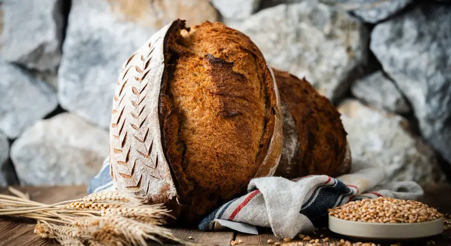 Zwei rustikale Brote, eines mit eingekerbtem Muster, liegen auf einem Holztisch. Weizenähren und eine Schale voller Körner deuten auf ein köstliches Ruchbrot Rezept hin.