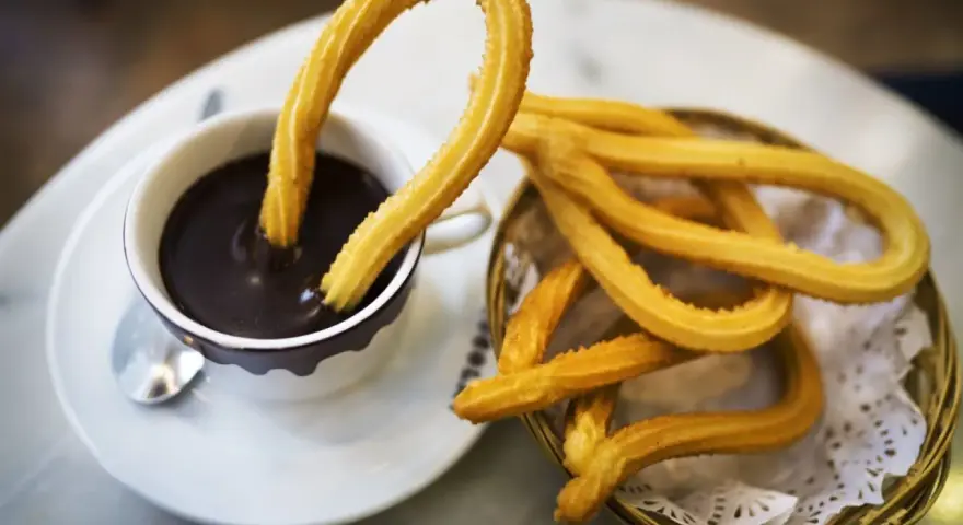 Churros sin gluten dorados y crujientes, uno sumergido en chocolate caliente, otro en una cesta.