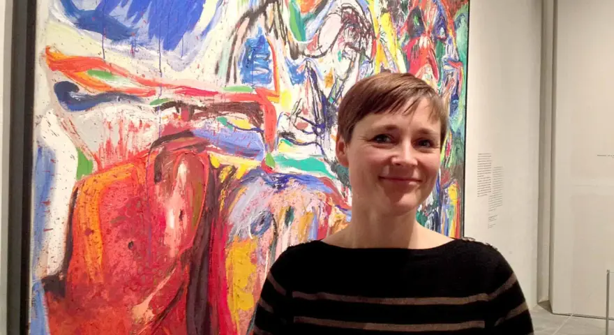 Kvinna ler framför en färgstark, abstrakt målning i Asger Jorns anda.