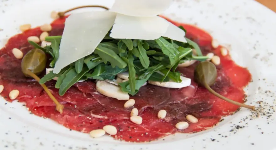 Delicato carpaccio di manzo con rucola, scaglie di parmigiano, pinoli, capperi e funghi.
