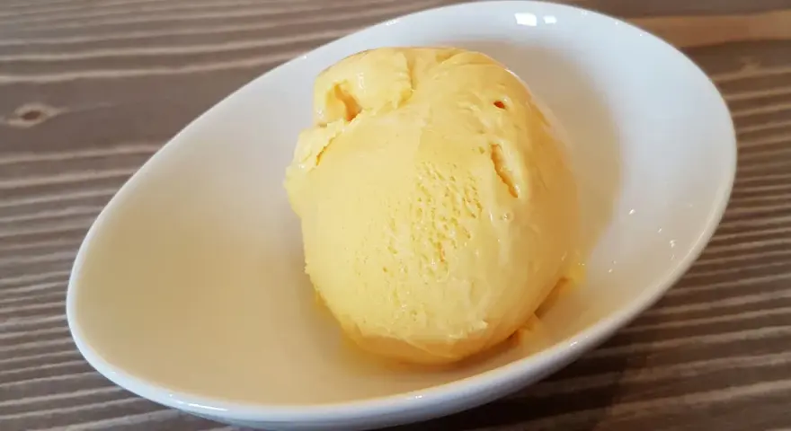 Ein Löffel cremiges, gelbes balla balla Eis in einer weißen Schale auf Holzhintergrund.
