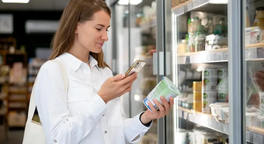Kobieta w białej koszuli trzyma jogurt i telefon, analizując produkt w sklepie.