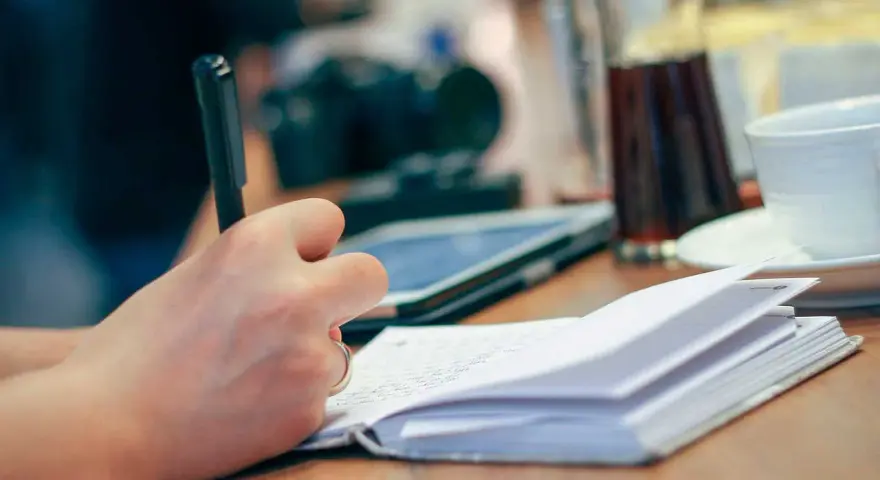 Mano escribiendo en un cuaderno abierto, inspirada para escribir cuentos. En la mesa hay una tableta, una taza y una jarra.