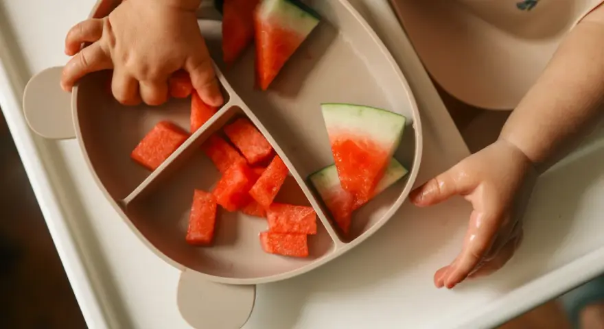Kleine Hände greifen nach saftigen Wassermelone-Stücken auf einem Teller. Ein süßer Wassermelone-Baby-Moment.