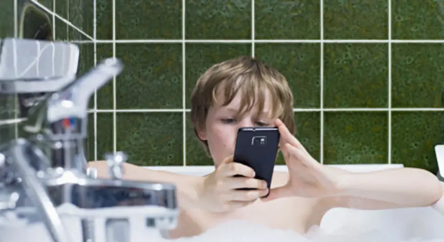 Chłopiec w wannie z telefonem. To jeden z objawów uzależnienia od telefonu u dzieci, gdy nawet kąpiel nie przerywa kontaktu z ekranem.