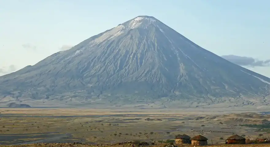 Ol Doinyo Lengai, wulkan w Tanzanii, góruje nad sawanną z kilkoma tradycyjnymi chatami Masajów.