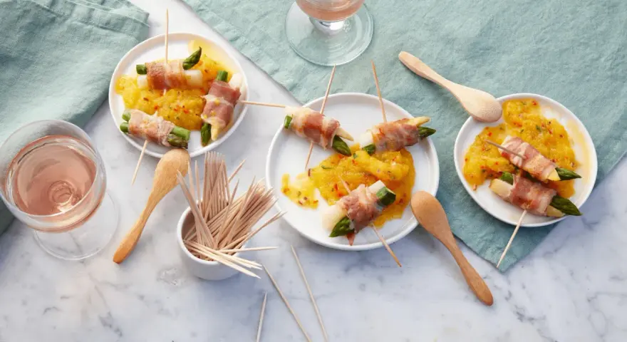 Leckere Spargel im Speckmantel auf gelbem Chutney, serviert auf kleinen Tellern mit Holzspießen und Roséwein.