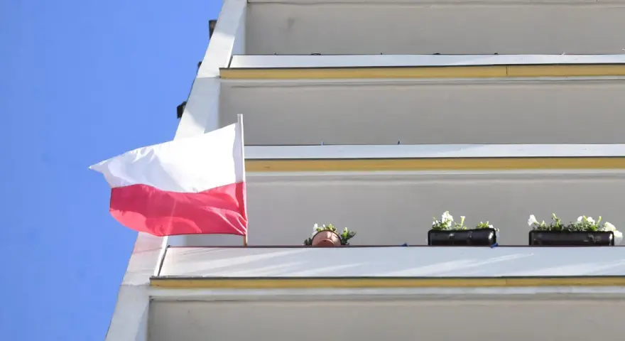 Prawidłowa flaga Polski powiewa na tle błękitnego nieba, obok balkonów z kwiatami.