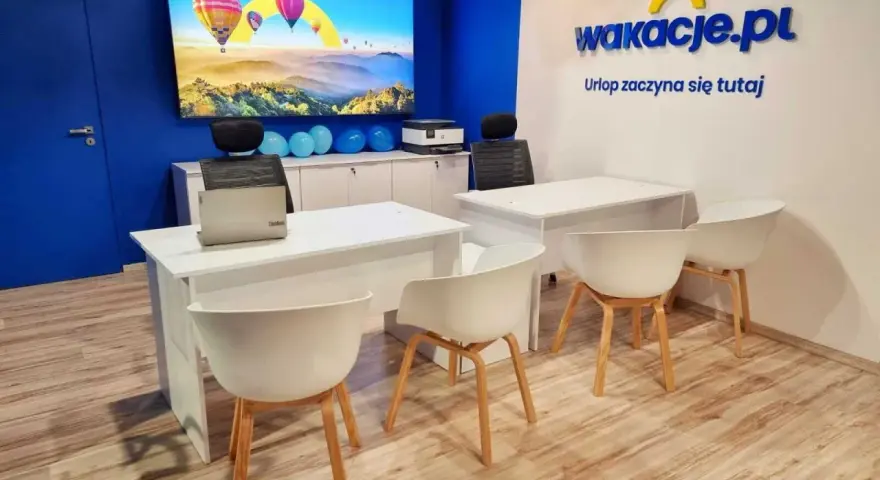 Nowoczesne biuro z logo "wakacje.pl", ekranem z balonami i białymi biurkami.