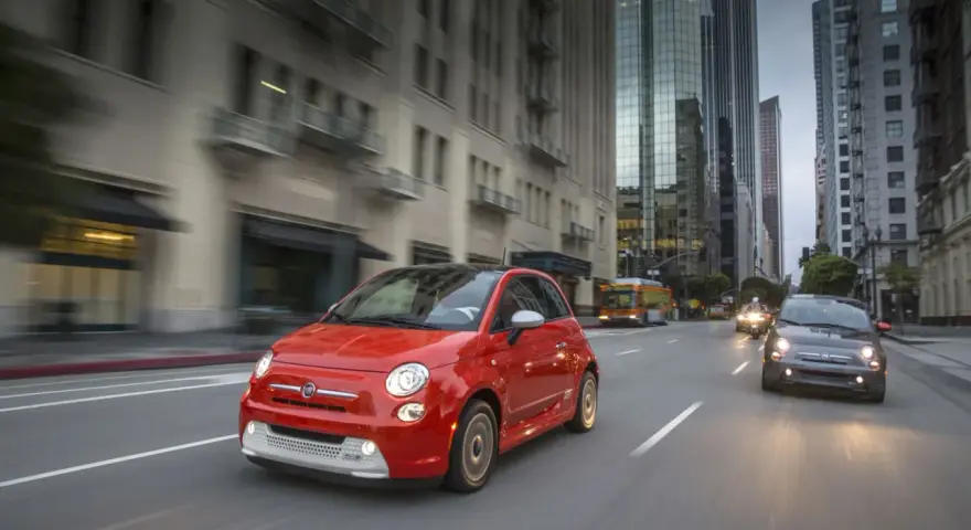 Czerwony Fiat 500e jedzie ulicą w mieście, w tle wieżowce i autobus.