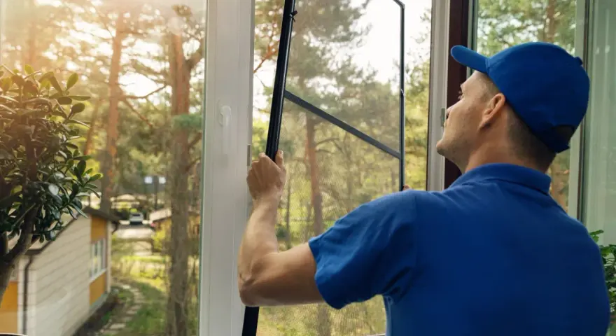 Man in blue shirt and cap performs window screen installation, preparing to fit the mesh into the frame.