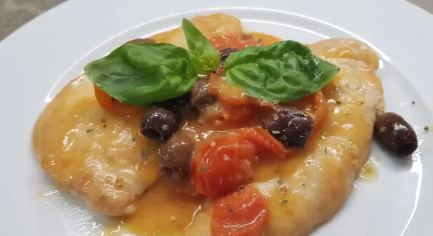 Delizioso pollo alla mediterranea con pomodorini, olive nere e basilico fresco su un piatto bianco.