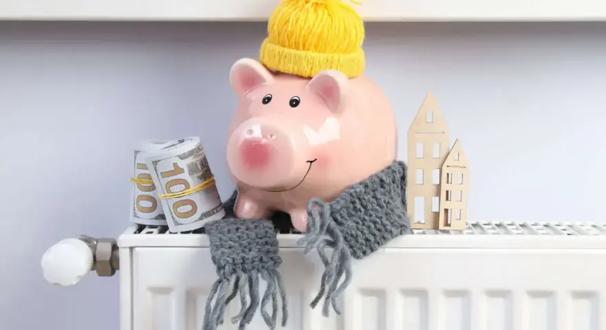 Świnka-skarbonka w czapce i szaliku, z banknotami i domkami na grzejniku. Oszczędzanie na rachunki za ogrzewanie, czyli bon ciepłowniczy w praktyce.
